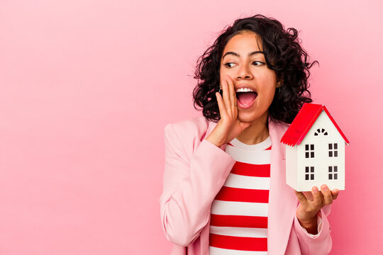Young latin woman holding a toy house isolated on pink background is saying a secret hot braking news and looking aside