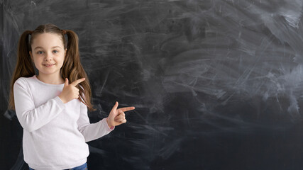 a Caucasian girl, a schoolgirl, is standing against the background of an empty chalk board. the index finger points to an empty space for text.