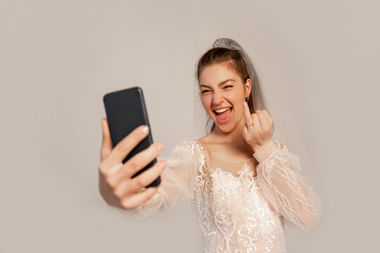 cheerful bride taking selfie on smartphone while showing wedding ring isolated on grey