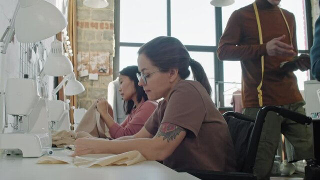 Medium shot with slowmo of seamstresses sitting at table and sewing garments in studio. Male fashion designer and his female assistant walking and discussing ideas