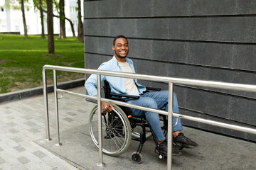 Happy impaired black man in wheelchair entering building on ramp outdoors, full length....