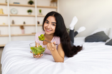Balanced nutrition concept. Lovely Indian woman eating fresh vegetable salad while lying on bed at...