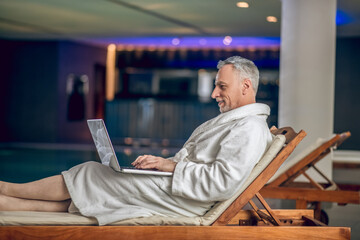 A gray-haired bearded man in a white bath robe spending time on internet