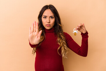 Young mexican pregnant woman holding pacifier isolated on beige background standing with...