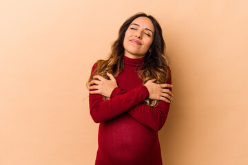 Young mexican pregnant woman isolated on beige background hugs, smiling carefree and happy.