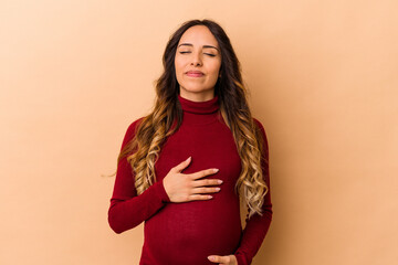 Young mexican pregnant woman isolated on beige background touches tummy, smiles gently, eating and satisfaction concept.