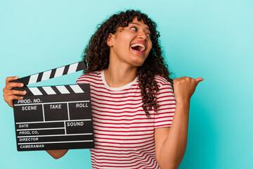 Young mixed race woman holding a clapperboard isolated on blue background points with thumb finger...
