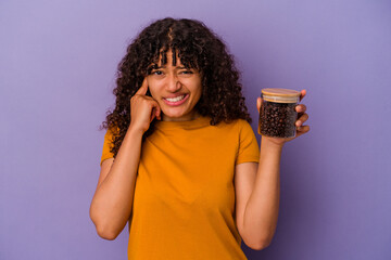 Young mixed race woman holding a coffee beans bottle isolated on purple background covering ears...