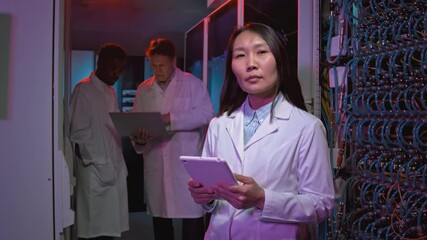 Tracking portrait with slowmo of female Asian network engineer in lab coat holding tablet and posing for camera in data center while her male colleagues with laptop discussing work - Powered by Adobe