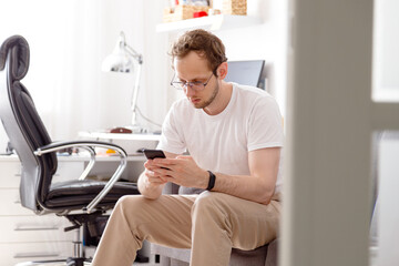 Happy young man in eyeglasses sitting on sofa sending text on mobile phone