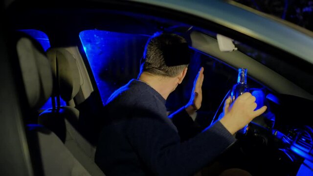 Booze addict automobile driver in black sweater raises hands with empty alcohol bottle in car salon stopped by police closeup