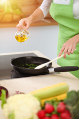 An attractive young dark-haired woman frying meal at worm pan by new keto recipe while standing and smiling in sunny kitchen. Cooking and householding concepts