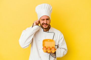Young caucasian chef man holding eggs isolated on yellow background covering ears with hands.