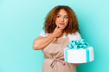 Young pastry latin woman holding a cake isolated on blue background looking sideways with doubtful and skeptical expression.