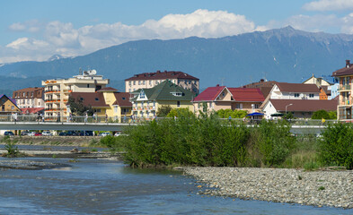 Pedestrian bridge over mountain river Mzymta against modern living buildings on Mzymta coastline nearby Sochi. Federal territory Sirius in Imeretinskaya lowland. Adler Sirius, Russia - May 18, 2021