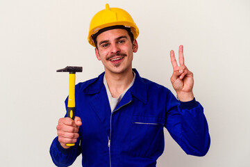 Young caucasian worker man holding a hammer isolated on white background joyful and carefree showing a peace symbol with fingers.