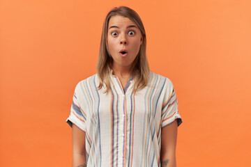 Surprised cute young woman in striped shirt looks amazed and looking at camera isolated over orange...