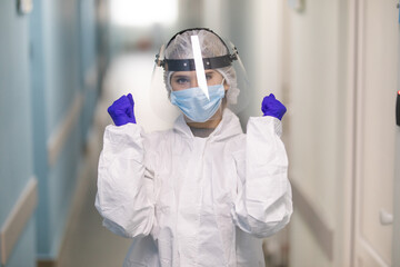 Closeup portrait of a young nurse with win gesture wearing a protective face shield and medical mask standing at the hospital corridor. 