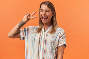 Cheerful attractive young woman in casual clothes winking and showing victory gesture isolated over orange background