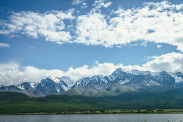 Awesome landscape with mountain lake and sunlit high snowy pinnacle among low clouds in blue sky. Atmospheric highland scenery with mountain lake under snow-white big mountain range in sunlight.