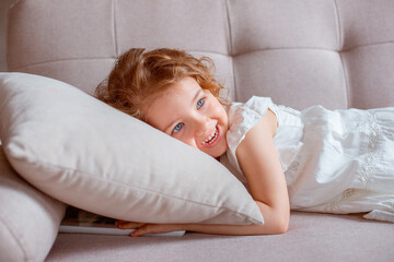 A happy sweet little girl is lying at home on the couch smiling, looking at the camera