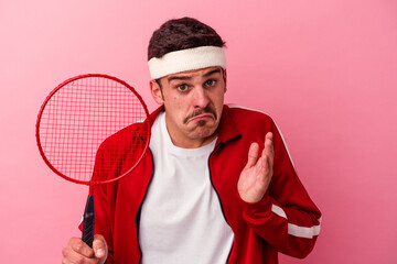 Young caucasian man playing badminton isolated on pink background shrugs shoulders and open eyes...