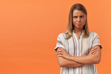 Thoughtful unhappy young woman in casual clothes thinking keeps arms crossed and looking away to the side at copyspace isolated over orange background