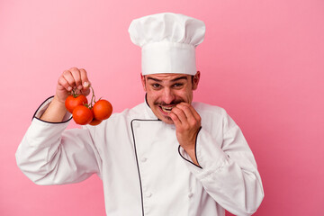 Young caucasian chef man holding tomatoes isolated on pink background biting fingernails, nervous and very anxious.
