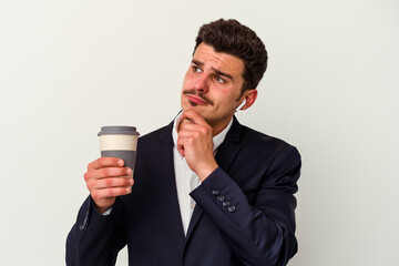 Young business caucasian man wearing wireless headphones and holding take way coffee isolated on white background looking sideways with doubtful and skeptical expression.