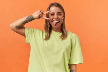 Smiling joyful young woman in yellow tshirt winking showing victory sign and tongue isolated over...