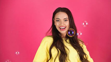positive young adult woman in yellow blouse with soap bubbles on pink background