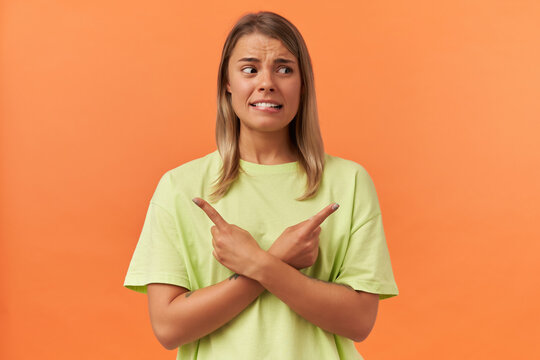 Confused Worried Young Woman In Yellow Tshirt Biting Lip And Pointing At Both Sides By Fingers Isolated Over Orange Background