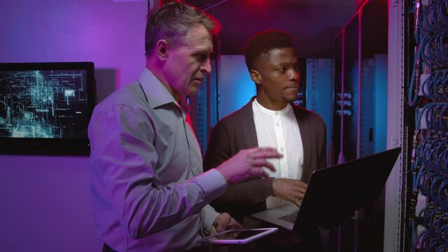 Tracking shot of middle-aged male Caucasian network engineer and his male African-American colleague holding laptop and tablet and talking while checking servers or switches in data center
