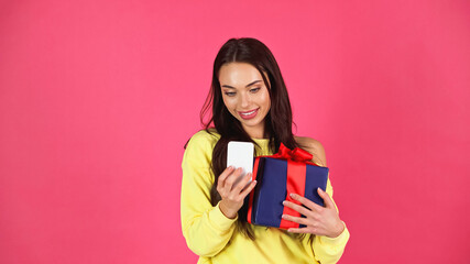 smiling young adult woman holding gift box and looking at cellphone isolated on pink