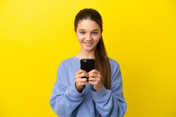 Fototapeta premium Little girl over isolated yellow background looking at the camera and smiling while using the mobile