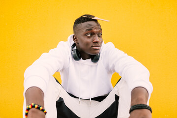 Young african American black man combing his afro hair while seated on a yellow wall background, wearing a white sweatshirt and wireless headphones. Looking to the side