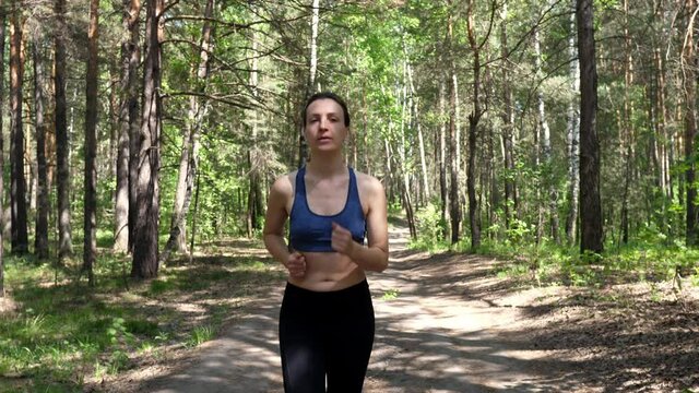 Sportive woman is engaged in fitness, runs on the road in a green park with trees. Running through the woods. Healthy body and mental health. Slow motion.