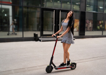 Cute, fit trendy girl on electric scooter driving through the city, looking at camera