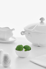 white porcelain dishware on a white background: soup tureen, gravy bowl, salt shaker and small plate with green broccoli , high key, shallow depth of field