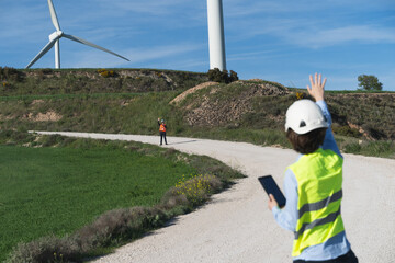 Focus on the worker in the orange vest. Unrecognized engineers wearing safety clothes working in wind turbine farm. They are making tests.