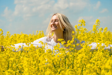 Fototapeta premium Happy smiling young blonde woman in a white shirt in a rapeseed field with her arms spread out to the side on a sunny day