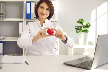 Important human organ. Portrait of a female cardiologist holding a little red heart while sitting at a desk in her office at the clinic. Concept of health, cardiology and heart disease prevention.