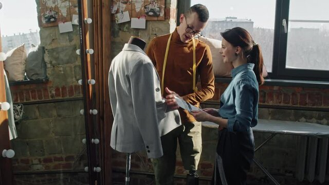 Dolly-in shot of male fashion designer with prosthetic leg inspecting jacket hanging on mannequin and talking to female seamstress showing him fabric samples in studio