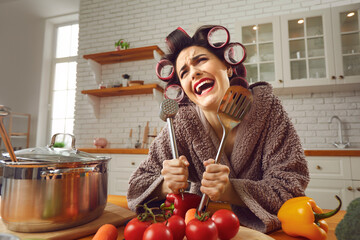 Funny crazy cheerful woman housewife in curlers and furry bathrobe standing in kitchen and holding...