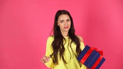 dissatisfied young adult woman holding gift box with gesturing with hand isolated on pink