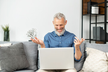 Modern cheerful senior man using laptop for video connection, mature grey haired businessman wearing casual shirt talking, looking into webcam, sitting on the comfortable couch at home