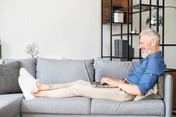 Optimistic middle-aged man lying on the couch and using laptop computer for messaging, happy...