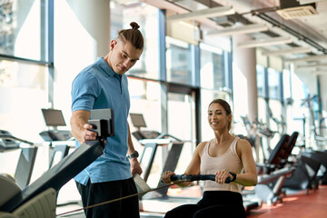 Young athletic woman using rowing machine while having sports training with male coach in a gym.