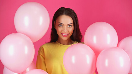 smiling young adult woman in yellow blouse among balloons isolated on pink