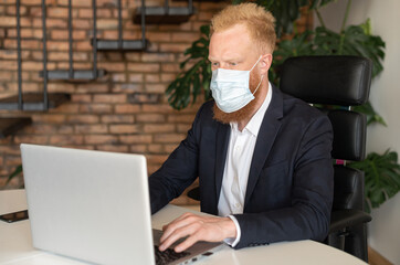 Serious red-haired businessman in smart casual suit wearing protective face mask using laptop computer sitting in modern office, male employee takes protective measures against viral disease